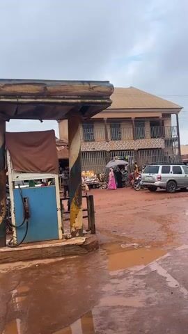 This video captures a bustling outdoor scene in Gembu, Nigeria. The foreground features a red, muddy, and wet surface, suggesting recent rain. A weathered gas pump, colored blue and yellow, stands prominently on the left, with a brown tarp draped over part of it.

Beyond the gas station, a marketplace unfolds. Numerous people are visible, some dressed in traditional African attire. Several motorcycles are parked, and a few are in motion. A person is riding a motorcycle with an umbrella, and another person is on the back, also with an umbrella. A white SUV is parked on the right side of the frame.

The background reveals a row of shops and buildings with corrugated metal roofs and some with awnings. The architecture appears functional and typical of a market area. The sky is overcast, indicating cloudy weather, possibly after rain. The overall atmosphere is lively and suggests daily commerce and activity. The presence of mud and puddles points to the wet season or recent rainfall. The time of day appears to be daytime, likely morning or afternoon, given the natural light. No visible text is present in the video.