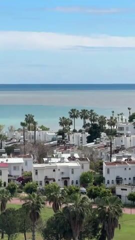 The video showcases a coastal cityscape with white-colored buildings, palm trees, and a visible shoreline. The sky is predominantly blue with scattered clouds. The water transitions from turquoise near the coast to a darker blue further out. The buildings are uniformly designed, with red-tiled roofs visible on some. Palm trees are interspersed among the buildings, adding a tropical feel. The environment gives the impression of a seaside town or resort.