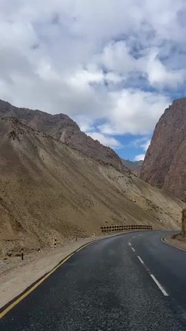 A narrow paved road winds through a mountainous landscape. The road is flanked by steep, rocky slopes on either side. Guardrails with black and yellow markings line the side of the road. The sky above is partly cloudy. The camera appears to be mounted on a moving vehicle, as the perspective shifts slightly throughout the clip. The overall environment suggests a remote, high-altitude region.