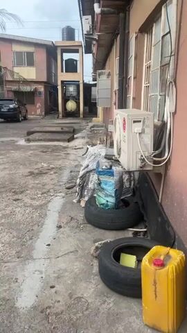 The video shows an outdoor, industrial-looking space with a building on the right. The building has a pinkish-tan exterior wall with multiple windows and an LG Dual Inverter air conditioning unit attached. Below the unit is a pile of items, including a black tire, a bag labeled "Bull", and a torn blanket. On top of the blanket, there is a small, broken wooden chair with a tabby cat peeking through the rungs. The cat appears to be staring directly at the camera. In the background, there's another building with a water tank structure. The ground is concrete and appears weathered and stained.