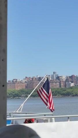 An American flag is attached to a pole on a boat. The flag is blowing in the wind. In the background, there is a body of water and a city skyline. The sky is blue and clear. The video appears to be taken from the deck of the boat, looking out over the water towards the city.