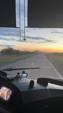 This media file captures a view from the dashboard of a Yutong bus as it travels along a highway in Botakara, Kazakhstan. The scene is set during either sunrise or sunset, indicated by the warm, orange and yellow hues illuminating the sky and casting a soft glow over the landscape. The sky is adorned with scattered clouds, adding depth and texture to the horizon.

The highway stretches ahead, a clear path leading towards a distant, conical mountain. The road is marked with white lines, suggesting a well-maintained route. On either side of the highway, the terrain is characterized by sparse vegetation, with some trees lining the left side of the road. The overall setting appears to be rural or semi-rural.

On the dashboard, several objects are visible. There's a GPS device mounted, a black knob, and a section of what appears to be a screen displaying images. The Yutong logo, a stylized circular design with the text "YUTONG" above it, is prominently displayed on the upper part of the windshield area.

The activity depicted is travel, with the bus in motion, moving away from the viewer and towards the setting or rising sun. The time of day, with its dramatic sky colors, evokes a sense of tranquility and the vastness of the open road. The image conveys a feeling of journey and perhaps a touch of peaceful solitude associated with long-distance travel. There are no people visible in the frame.