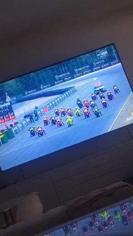 A television screen displays a motorcycle race. A pack of motorcycles is visible on a blue track with yellow lines. Several motorcycles are in the foreground, with more spread out behind them. A white vehicle and a few additional motorcycles are further down the track. The screen shows sponsorship logos from "Monster Energy". The environment surrounding the television is dimly lit, with furniture and a blanket visible in the lower portion of the frame. The camera is positioned at an angle, capturing the screen and its surroundings.