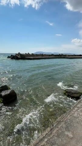 The video shows a body of water with waves gently lapping at the shore. The water is a clear blue-green, and the sun is shining, creating sparkles on the surface. There are several large rocks in the foreground, some of which are covered in green algae. In the background, there is a concrete breakwater with several large, angular concrete blocks forming a barrier against the waves. Beyond the breakwater, the ocean stretches to the horizon, where a faint, hazy outline of land can be seen. The sky is a bright blue with a few white, fluffy clouds. The overall scene is peaceful and serene, evoking a sense of calm and tranquility.