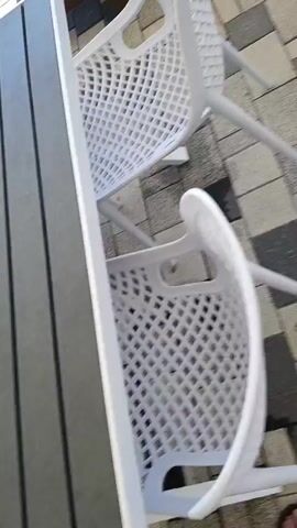 The video pans across an outdoor patio area. The patio is paved with light and dark square tiles arranged in a patterned fashion. To the left, a white plastic chair is visible. In the background, there is a wooden bench with blue cushions and a dark-colored table with a dark metal base. Beyond the patio furniture, a railing overlooks a beach with white umbrellas. The sea is visible in the distance, with several people swimming and two surfboards standing upright. The sky is clear and blue. The video then pans to the right, showing more patio furniture and the sea. A few people are visible in the water, and a jet ski is in the distance. Another table and chairs are visible in the foreground. The video continues to pan, showing more of the patio and the sea, with some water attractions visible in the distance.