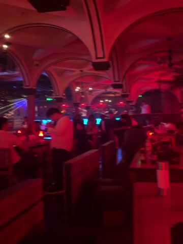 The interior of a bar or club is depicted in the video. The space is dimly lit with red and blue lighting, and the architecture features arches with black trim. People are milling about, some sitting at tables, others walking through the space. There are candles on the tables, and liquor bottles are visible in the foreground. The lighting creates a club-like atmosphere. The camera appears to be moving slightly as it captures the scene. There is music playing.
