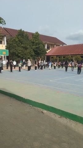 This media file shows a group of people, likely students, participating in what appears to be a marching band or parade practice on a brightly colored outdoor court. The setting is a school or community complex, with a long, multi-story building with a reddish-brown tiled roof dominating the left side of the frame. Lush green trees are interspersed around the building and on the court. The court itself is marked with various colored lines, suggesting it could be a basketball or volleyball court.

The subjects are organized in neat rows, facing forward. Many are holding instruments, with at least two individuals in the foreground holding large bass drums. Others appear to be holding flags, some of which are colorful, and one prominent flag in the middle of the frame is red and white, resembling the Indonesian flag. Some individuals are wearing uniforms, while others are in casual attire. The overall impression is one of organized activity and practice.

The time of day appears to be daytime, with overcast or cloudy skies contributing to diffused lighting. There are no strong shadows, and the colors of the court and building are clearly visible. The activity seems to be a rehearsal, possibly for an upcoming event. The participants appear focused and engaged in their tasks.

The location cues, specifically the Indonesian flag, strongly suggest the event is taking place in Indonesia, as noted by the provided location context of Rhieng Blang. There is no visible text in the media file.