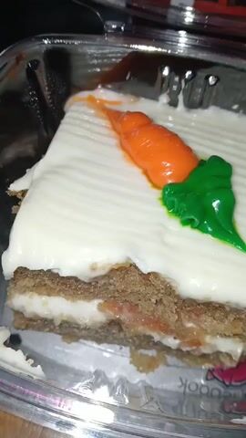 A close-up video shows a slice of carrot cake in a clear plastic container. The cake has two layers of brown cake with a white cream filling in between. The top of the cake is covered in a thick layer of white frosting with horizontal lines and is decorated with a small orange carrot and a green leaf, both likely made of frosting or fondant. The camera moves slightly around the container to show the side of the cake.
