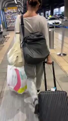 A woman walks away from the camera at an airport, dragging a rolling suitcase with her right hand. A large shoulder bag is on her left shoulder, and a colorful shopping bag is in her left hand. The airport terminal has benches, newsstands, and taxis parked on the street outside. The woman is wearing a light-colored top and light-colored pants.