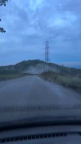 The video, shot from the perspective of a vehicle's dashboard, shows a road leading towards a hilly, mist-shrouded landscape under a cloudy blue sky. A large electricity pylon stands prominently on the right side of the road, with power lines stretching across the scene. Additional smaller utility poles are visible on the left side. The road appears unpaved or poorly maintained, with a rough, dark surface. As the video progresses, the vehicle moves forward, showing more of the road and the surrounding mist and greenery.