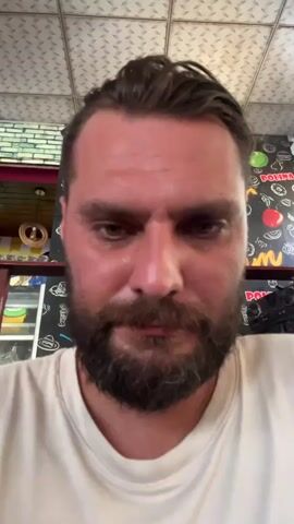 A close-up video shows a man with a beard and dark hair, looking directly at the camera. He is wearing a light-colored t-shirt. The background is blurred, but appears to be an indoor setting with a ceiling that has a tiled pattern. There are some objects and decorations visible behind him, including a TV screen displaying a sunset, and what looks like food items on a counter. The man speaks in a language that appears to be Russian. The video cuts to a wider shot where a person is visible sitting at a table, drinking from a cup. The overall environment seems to be a casual dining area.