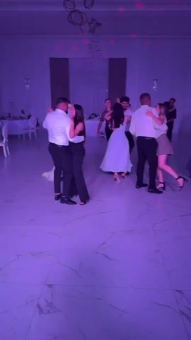 The video shows a group of people dancing in a well-lit room. The room appears to be a banquet hall or ballroom, with a polished marble floor. There are several couples dancing, and others standing around watching. The lighting is varied, with pink and green spotlights creating a festive atmosphere. In the background, there are tables set with white cloths and chairs, suggesting a formal event such as a wedding reception or party. The people are dressed in formal attire. The camera is moving slightly, panning across the scene and capturing the interactions of the dancers.