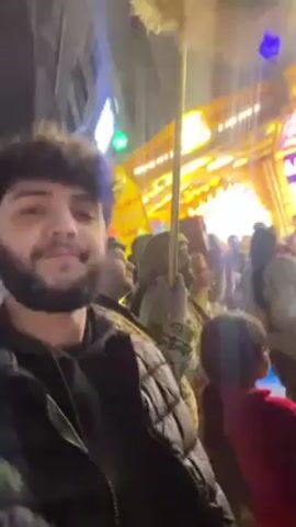 The video shows a crowd of people gathered outdoors at night. Many are holding and waving flags. A man with curly hair and a beard, wearing a dark quilted jacket, appears to be taking a video with his phone, facing the camera at times. Children are visible in the crowd. The background is filled with lit-up storefronts and buildings. The crowd is moving and engaged in some form of event or celebration.