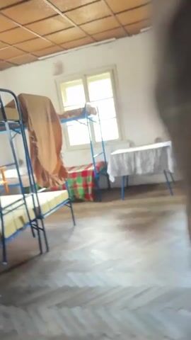 The video pans across a dimly lit, sparsely furnished room that appears to be a dormitory. There are at least two sets of blue metal bunk beds, with yellow mattresses and some bedding visible. A table covered with a patterned cloth stands against a white wall, with a blue base. Two windows with beige frames are present. The floor is made of wooden planks arranged in a herringbone pattern. The ceiling is made of brown textured panels. A chest of drawers with white fronts is visible in the background. The room appears to be unoccupied.