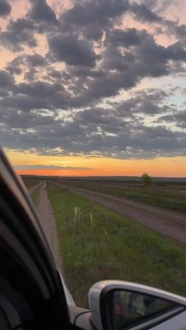 The view from inside a moving car shows a sunset landscape. The car travels along a dirt road, flanked by green fields. The sky is filled with textured grey and white clouds, illuminated by a vibrant orange sunset. A single, small tree is visible on the horizon. The car's side mirror offers a blurred reflection of the scene.