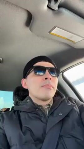 Here's a description of the video:

The video shows a man in a car, presumably driving. He is wearing a black beanie, sunglasses, a patterned shirt, and a black puffer jacket. The camera angle is from below, looking slightly up at him. The interior of the car is visible, including the sun visor. Music is playing in the background.