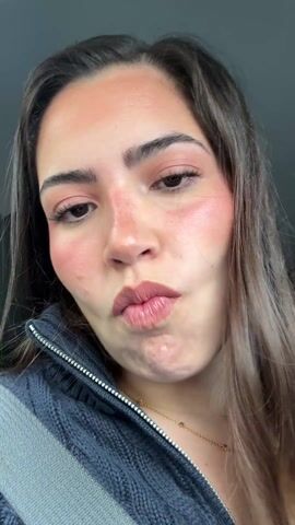 A close-up video shows a woman with light brown hair and fair skin making various faces at the camera. She is wearing a navy blue knit sweater with a silver zipper and a delicate gold necklace. Her makeup includes noticeable blush on her cheeks and a pink lip color. The background is dark, suggesting she may be inside a car. She puckers her lips, winks, and makes other expressions, all while looking directly at the camera. There is faint audio in the background, possibly music or conversation.