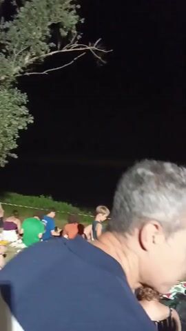 The video shows a gathering of people sitting on a grassy area at night, facing a body of water. In the foreground, a man with graying hair and a dark shirt is looking to the right. In the background, numerous individuals are seated on blankets and chairs, some appearing to be watching something across the water. A faint, long structure, possibly a bridge or a pier, is visible spanning the water in the distance, with lights along its length. The sky is dark, indicating nighttime.
