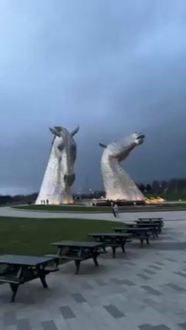 The video shows two large, silver horse head sculptures in a park-like setting under a cloudy, overcast sky. The sculptures are positioned on a grassy area with lights around the base. Several picnic tables are arranged in a row on a paved area in the foreground. Two people are walking on the paved area towards the sculptures. The overall lighting suggests it is dusk or early evening.