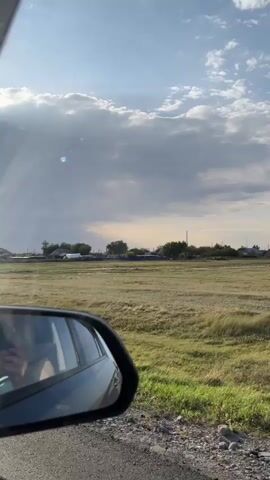 The video shows a view from inside a car, looking out the windshield and side mirror. The sky is partly cloudy with the sun shining brightly. In the distance, there appears to be a small town or village with some buildings and trees. The car is driving on a dirt road with dry grass on either side. The windshield is dirty with a number of smudges and streaks. The side mirror shows a reflection of a person’s face.