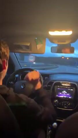 A person is driving a car with music playing. The person has short brown hair, wearing a dark hoodie, and is seated in the driver's seat with their hands on the steering wheel. The car appears to be driving on a highway during the late evening or early morning based on the light conditions. The car's dashboard is visible, showing the speedometer reading 84 and a touchscreen displaying music information.  Outside the car, there are other vehicles on the road and a landscape with hills.