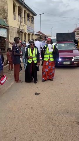 Two women walk in front of a crowd, each wearing a high-visibility vest over a traditional outfit. The woman on the left wears a black garment with a maroon trim and sunglasses. The woman on the right holds a microphone and a stack of papers. She is wearing a red and orange patterned dress and a headscarf. Behind them, a banner with the letters "EP" is visible. People are walking behind the women, some are looking forward, some are looking at the women. The scene appears to be outdoors on a street with buildings on either side. There is a car with its doors open in the background. The ground is paved and there is a piece of debris on the ground.
