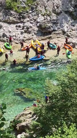 The video shows a group of people on the rocky bank of a clear, turquoise river. Several inflatable rafts, predominantly yellow and some smaller green and blue kayaks, are pulled up onto the shore or in the shallow water. People wearing helmets and life vests, dressed in dark red or blue wetsuits, are gathered around the boats, some appearing to be preparing for an activity or having just finished. A few individuals are in the river itself, either swimming or paddling in a kayak. The river flows through a gorge with rocky, steep sides covered in sparse vegetation and trees. The sun is shining, casting shadows and highlighting the water's clarity. The overall scene depicts a recreational activity, likely whitewater rafting or kayaking, in a natural, outdoor setting.