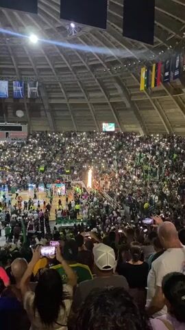 The video shows an aerial view of a basketball game in a large, domed stadium. The stands are filled with spectators, many of whom are holding up their phones with the flashlights on, creating a sea of lights. On the court, players are gathered around the center, and there are fireworks or pyrotechnics being set off. The atmosphere appears festive and celebratory, with many people taking photos or videos of the event. Flags are visible hanging from the ceiling of the stadium.