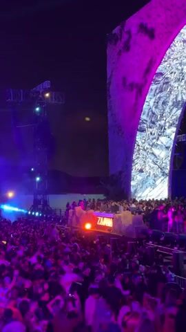 The video shows a crowded outdoor concert at night. A DJ booth labeled "ZAMNA" is elevated in the center. The crowd is large and dense, mostly facing the stage. Stage lighting and lasers illuminate the crowd and the stage area. Behind the DJ booth is a large decorative arch structure with marble-like texture and pink lighting on one side. A lighting tower is positioned on the left side of the stage. The sky is dark, and the overall atmosphere is vibrant and energetic.
