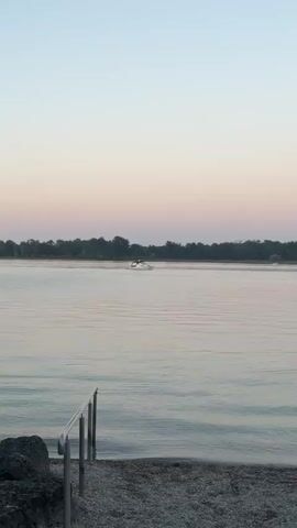 The video shows a tranquil scene of a body of water, likely a lake or river, at dusk. The sky displays a soft gradient of pale blue and pink hues. In the foreground, a gravelly shore is visible, with a metal railing leading down towards the water. The water itself is calm, with gentle ripples reflecting the sky's colors. A small white boat is moving across the water in the mid-distance, leaving a faint wake. Further in the distance, a line of trees forms the shoreline. A duck is briefly seen swimming on the water.
