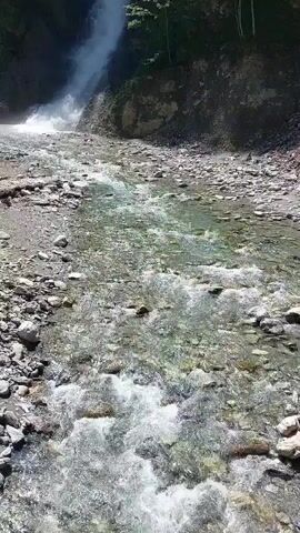 The video shows a waterfall cascading down a rocky cliff face into a shallow, clear stream. The stream flows over a bed of pebbles and rocks. Trees and vegetation grow on the slopes of the cliff. The overall scene depicts a natural, outdoor environment.