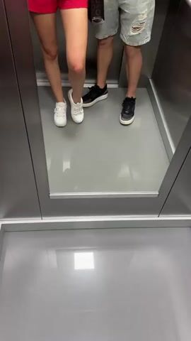 The video shows a couple in an elevator. The woman is on the left wearing red shorts and a black lace top, holding a black handbag with a gold chain. She is also holding a smartphone and taking a selfie. The man is on the right wearing light blue ripped shorts and a beige t-shirt. He is holding a smartphone. The elevator has stainless steel walls and a grey floor. There is a mirror on the back wall reflecting the couple. The floor of the elevator has a bright square light. The video is shot from a low angle looking upwards.