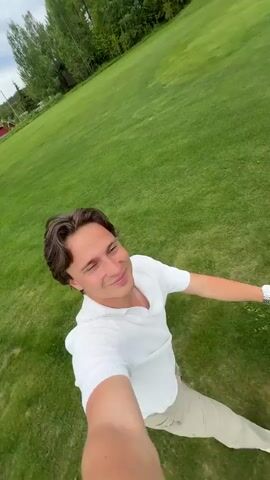 The video showcases a person playing golf. The first shot is from a high angle, showing a man with short brown hair, wearing a white long-sleeved polo shirt and tan pants. He is looking off to the side.  Next, the camera angle changes to show a golf cart on a green golf course. Two people are seen walking in the background. The focus then shifts to the golf cart's handle, and a person pushes the cart forward. The cart holds a golf bag filled with clubs and various hats, including a light green one with an emblem and a blue one. The environment includes a vast, green golf course with rolling hills and trees in the background under a cloudy sky.