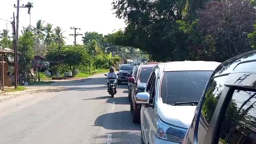 The video captures a street scene with moderate traffic. On the right side of the road, several cars are parked or moving slowly in a line. These include a silver van, a white minivan, and a black car, all partially visible. A red truck with an open cargo bed is moving on the road, its driver visible in the cab. On the left side of the road, a motorcycle with a rider wearing a helmet is moving forward. There are also some small buildings and palm trees along the roadside, suggesting a rural or semi-urban environment. Power lines and poles are visible in the background. The lighting indicates it is daytime, likely sunny.