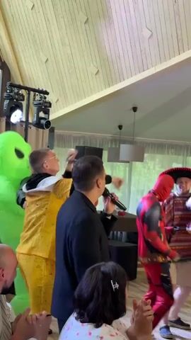A video shows a group of people in costumes dancing and interacting at an indoor event. On the left, a person in a yellow and green costume with large hands gestures upwards, appearing to be in mid-dance. To their right, a person in a red and black Deadpool costume faces away from the camera, with their arms raised. Next to Deadpool, a person in a dinosaur costume with an open mouth is visible. In the background, a person in a colorful striped shirt and a wide-brimmed hat stands near a table with a microphone. The environment appears to be a room with wooden-paneled walls and a slanted ceiling, with artificial lighting fixtures and decorative elements like lamps and curtains. Other guests are seated or standing, observing the activity. The overall atmosphere suggests a lively celebration or party.
