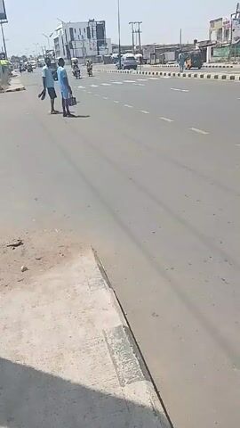 The video shows a daytime scene on a paved road. Two men in light blue shirts and dark shorts stand on the median of a multi-lane road. They then cross the road, joined by a third man in an orange shirt and shorts. They walk in front of passing cars. Other traffic including motorcycles and auto rickshaws are visible. Billboards and buildings line the road. The video is taken from a slightly elevated position, likely from the side of the road.
