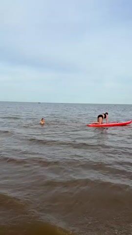 This video captures a serene moment at a beach in Ondvor, Russia. The scene is set on a calm body of water, likely a lake or a bay, with a gentle wave pattern rolling towards the shore. The sky is overcast with a pale, diffused light, suggesting it's either early morning or late afternoon on a cloudy day.

The main subjects are two individuals in the water. In the foreground, a person is on an orange paddleboard, positioned in a kneeling stance. They are wearing dark-colored swimwear. Further back and to the left, another person is swimming, with only their head and shoulders visible above the water. They appear to be a child or a young person. The presence of two small boats on the horizon hints at recreational activity or local transport in the distance.

The activity is leisurely water recreation. The person on the paddleboard seems to be either resting, preparing to paddle, or observing the swimmer. The swimmer is enjoying the water, possibly playing or exercising. The overall mood is peaceful and relaxed, typical of a calm day at a natural water body. The shoreline is visible on the far left, showing wet sand and some sparse green vegetation, grounding the scene in a beach environment. There is no visible text in the media.