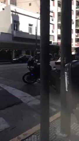The video shows a city street on a sunny day.  A black motorcycle is parked on the sidewalk. Cars drive by, including black taxis. A bus drives through an intersection.  Buildings line the street, with businesses visible at ground level.  People walk on the sidewalks and crosswalks. The camera moves and pans across the scene.
