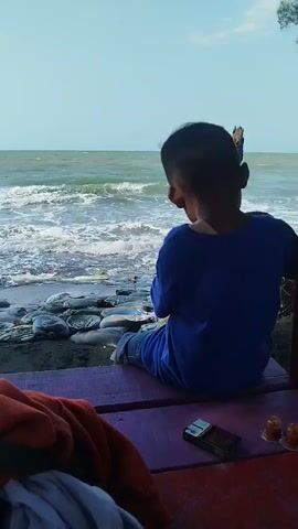 A boy in a blue shirt sits on a purple bench facing the ocean. The waves are crashing on the shore. There are several white objects, possibly bags of sand or debris, on the beach in front of the boy. On the bench next to the boy, there is a cigarette lighter and two small orange cups. The sky is a clear blue.