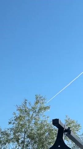 A contrail stretches diagonally across a clear blue sky. The contrail appears white and wispy, indicating it is likely from an aircraft. In the lower left of the frame, the top branches of a green-leafed tree are visible, partially obscuring the sky. A dark, angular object, possibly a camera mount or tripod leg, is also present in the lower right corner. The contrail starts at the upper edge of the frame on the right and extends towards the left, eventually disappearing behind the tree. The scene is brightly lit, suggesting daylight.