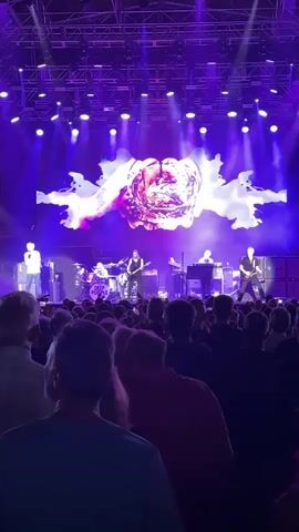 The video shows a live concert performance by the band Deep Purple. A vocalist stands center stage, singing into a microphone. To his left, a drummer plays a drum kit adorned with the band's name. A guitarist is positioned center-right, actively playing a guitar. A keyboardist is seated on the right, also playing an instrument. The stage is illuminated by bright purple lights, and a large screen behind the band displays abstract, colorful visuals. A crowd of concertgoers stands in the foreground, facing the stage, with many holding up their phones to record the performance. The overall atmosphere is energetic and engaging.