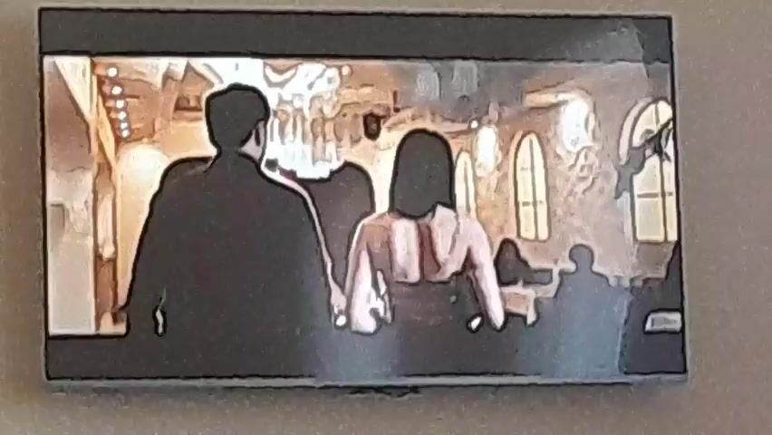 The video displays a television screen showing an animated or filtered scene set indoors. Two central figures, a man with dark hair and a beard, and a woman with dark hair, are initially seated at a table. The man wears dark long sleeves, and the woman wears a dark, possibly sleeveless, top. The background features light-colored, textured walls and arched windows. Initially, both individuals are partially facing away from the viewer, then turn to show their profiles. Subsequently, the man stands up from his seat and moves behind the seated woman, placing his hands on her shoulders or upper arms. The woman tilts her head back slightly as the man stands behind her. Other blurred figures are visible in the deeper background.