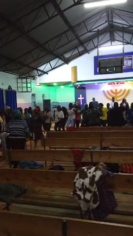 The video shows a group of people dancing in a church. The interior of the church has wooden pews in rows. Some blankets and clothes are on the pews. The stage has a white cross on either side of a golden menorah. There is also a television hanging above the stage. The people are dancing facing the stage. The lighting is varied, with green, blue, and purple lights on the stage. Some people are dressed casually. The church has a metal ceiling and appears to be well lit.
