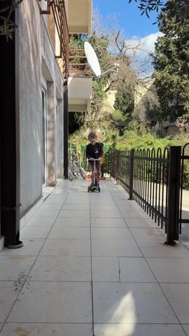 A young boy, approximately 5 years old, rides a small, green scooter toward the camera on a tiled patio. He is wearing a long-sleeved black shirt and brown pants. The patio is bordered on one side by a building with sliding glass doors and a balcony with a satellite dish. On the other side, a black metal fence separates the patio from a green, overgrown yard with trees and bushes. Bicycles are visible in the background. The sky is blue with some scattered clouds. The boy stops the scooter and looks directly at the camera.