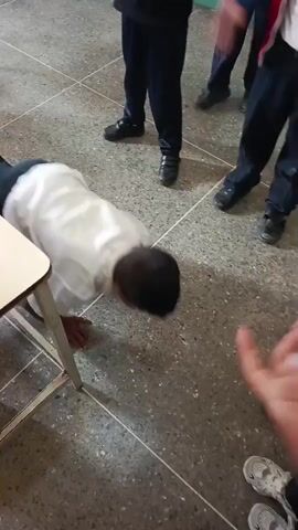 A child in a white shirt and dark pants is on the floor performing push-ups. Other children in similar attire are standing around watching and seemingly counting. The floor is made of grey tiles with white lines. A white table is visible to the left of the child performing push-ups.