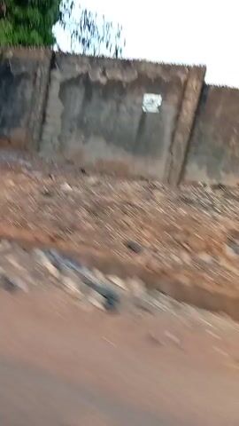 The video shows a moving perspective from a vehicle traveling along a paved road. The scenery consists of dilapidated buildings made of metal sheets and concrete walls with visible wear and tear. The environment appears to be a low-income area with a somewhat dusty and neglected look. There are no people visible in the frame, and the primary focus remains on the structures and the road as the vehicle passes by. The sky is partially visible, indicating it is daytime. The footage seems to be taken from the passenger seat or a similar vantage point.