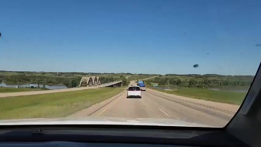 The video shows a first-person perspective from inside a vehicle driving on a highway. The sky is clear and blue. A bridge with multiple arches is visible to the left, spanning over a body of water. In front of the vehicle, on the highway, there are other vehicles, including a white van and a blue truck. The surrounding landscape is green with trees and fields. The vehicle appears to be moving at a moderate speed.