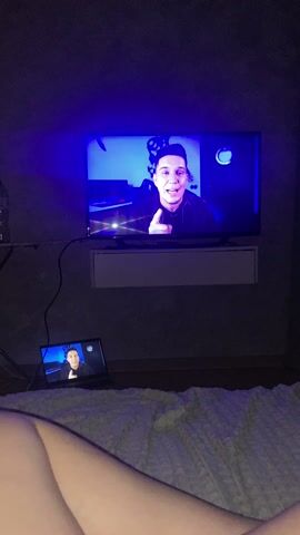 A person is lying on a bed in a dimly lit room, watching a video on a TV and a laptop simultaneously. The TV is mounted on a wall with a small cabinet underneath. The video being watched on both screens features a man speaking directly to the camera. The bed is covered with a white textured blanket. The walls are a dark grey color. The room has a blue-ish hue from the TV and laptop screens. The laptop is open on the bed near the person. The video switches off abruptly.