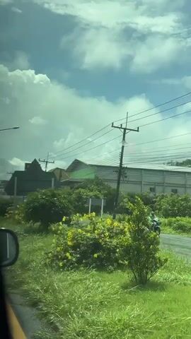 The video shows a view from inside a vehicle, looking out at a roadside scene. The road appears wet, suggesting it has recently rained. Lush green grass and vegetation line the side of the road. Several utility poles with electrical wires are visible. In the background, there are buildings, including a yellow building with a prominent sign that reads "JOTUN". A black pickup truck is parked on the right side of the road. The sky is partly cloudy with white clouds against a blue sky. The overall impression is a view of a suburban or semi-rural area on a cloudy day.