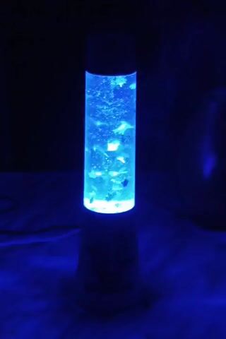 The video shows a blue lava lamp with small objects resembling fish floating in a clear liquid. The lamp is cylindrical and emits a bright blue light from the base, illuminating the contents. The background is dark, making the lamp the central focus. The objects within the lamp appear to be moving slowly, creating a calming visual effect. There is a cable visible on the left side of the lamp, suggesting it is powered on. The lamp sits on a dark surface that reflects the blue light.